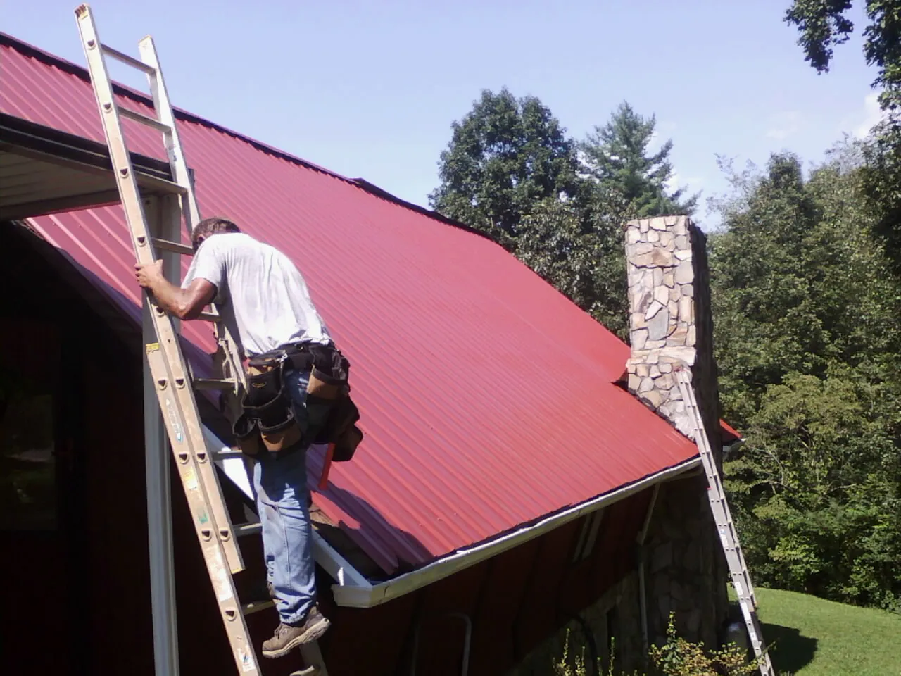 Roofer on a ladder beside a red metal roof and stone chimney — Soffit & Fascia Repair by Roofing Brothers in South Beloit
