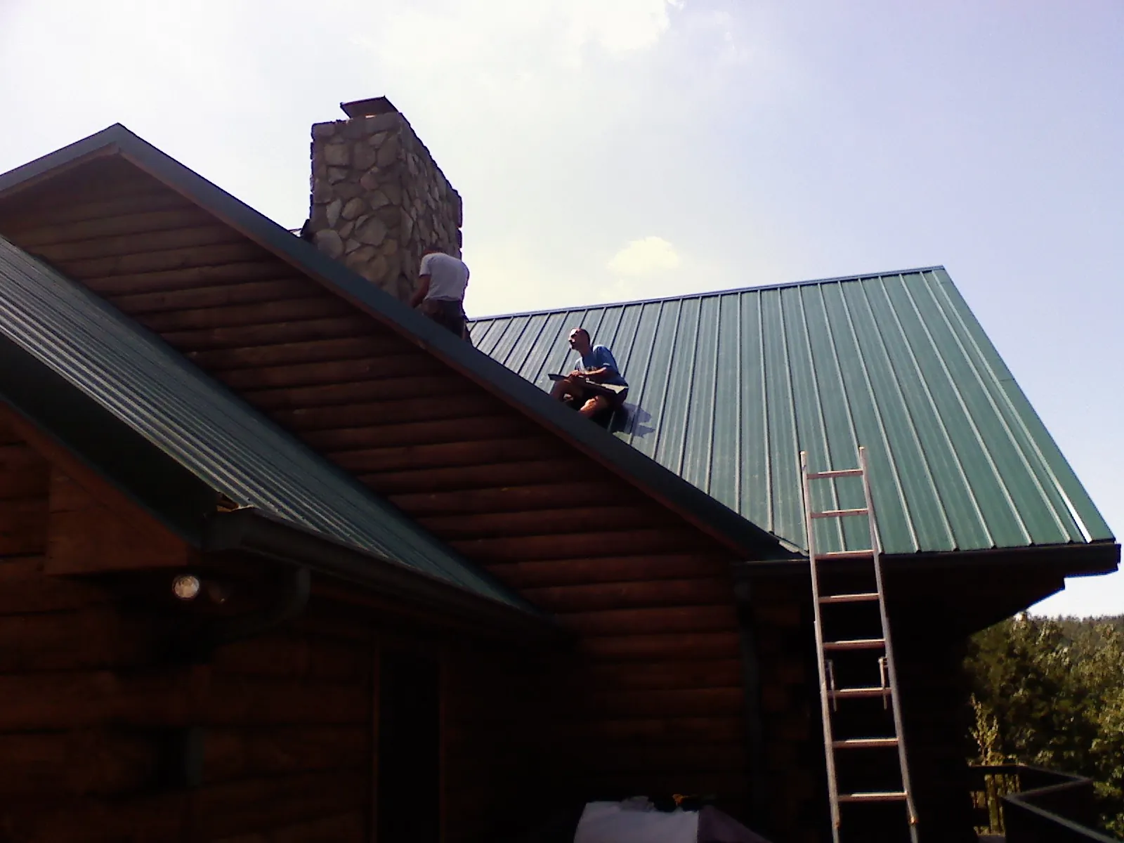 Roofers installing green metal panels beside a stone chimney on a log home in South Beloit